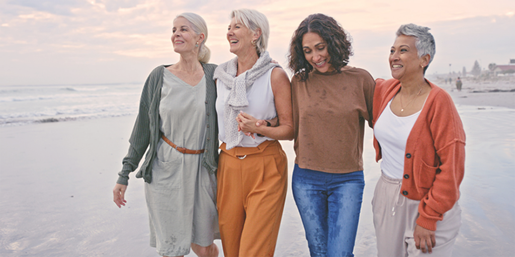 Four women walk along the beach, laughing and smiling.