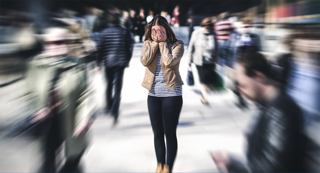 A girl with social anxiety disorder stands with her hands covering her eyes while panicking in a crowded public space during the middle of the day.