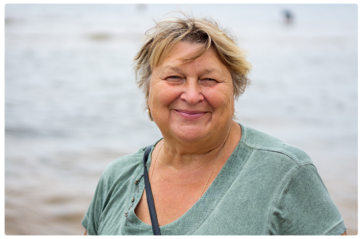 An overweight woman in a green shirt smiles while standing on the beach on an overcast day.